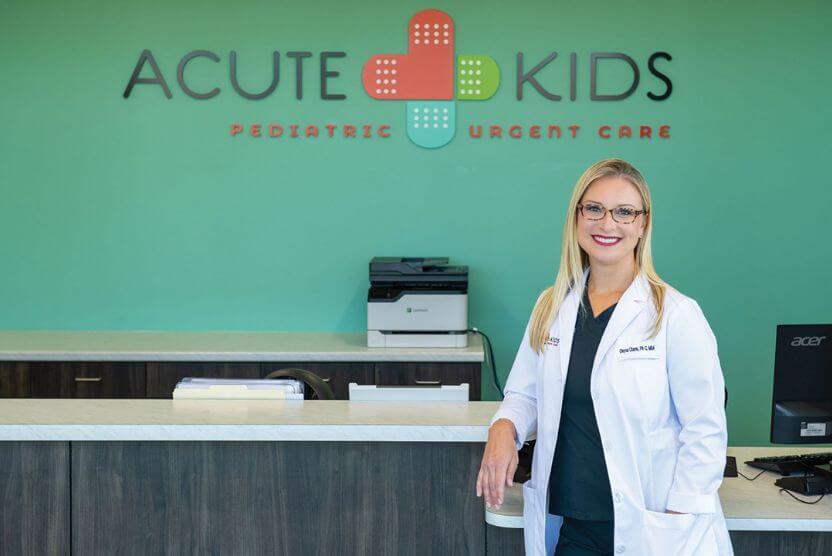 Chrysa Charno, dressed in a medical coat, standing in front of the AcuteKids Urgent Care sign.