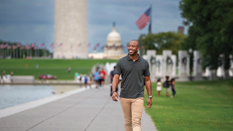 RIT Alum Arthur Deane is wearing a greay polo short and beige pants. He walks along a path in Washington, D.C., with the U.S. Capital Building in the distance. 