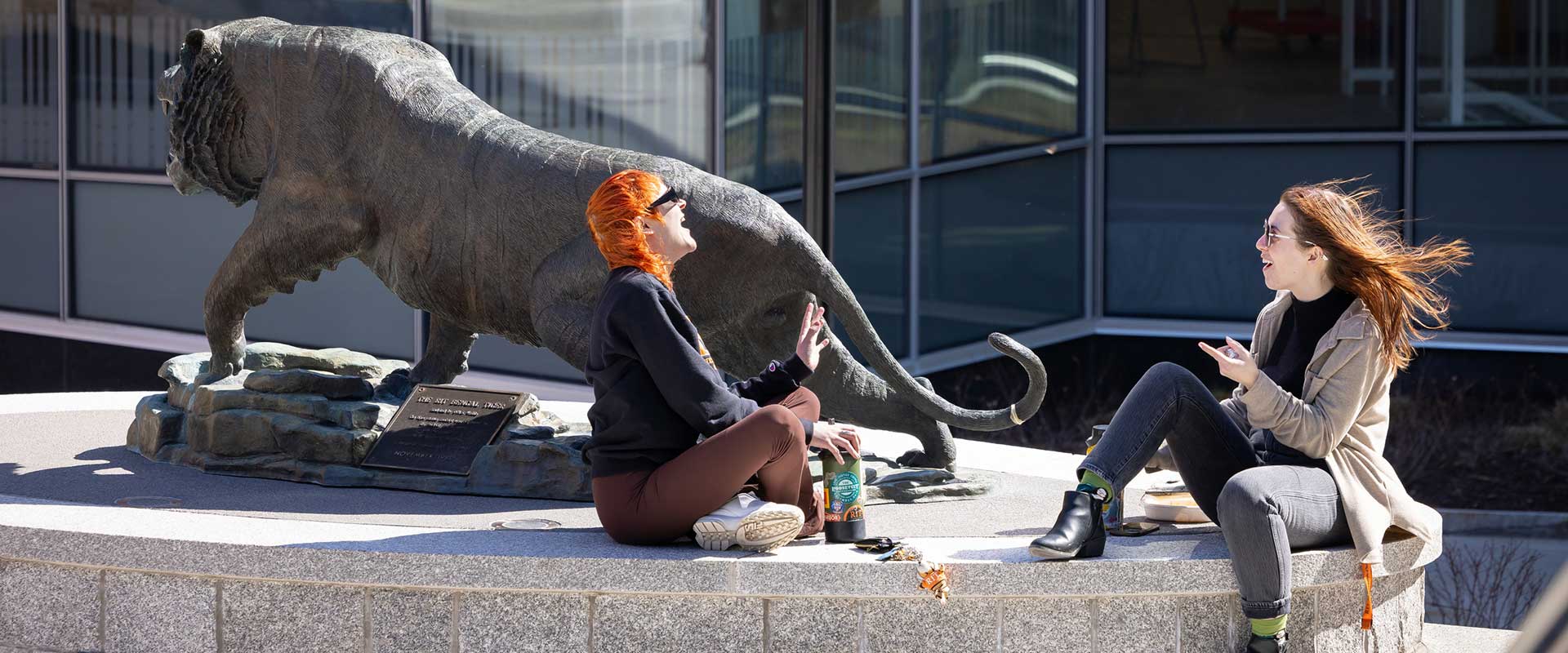 Two students sitting by a tiger statue, having a conversation and laughing on a sunny day.
