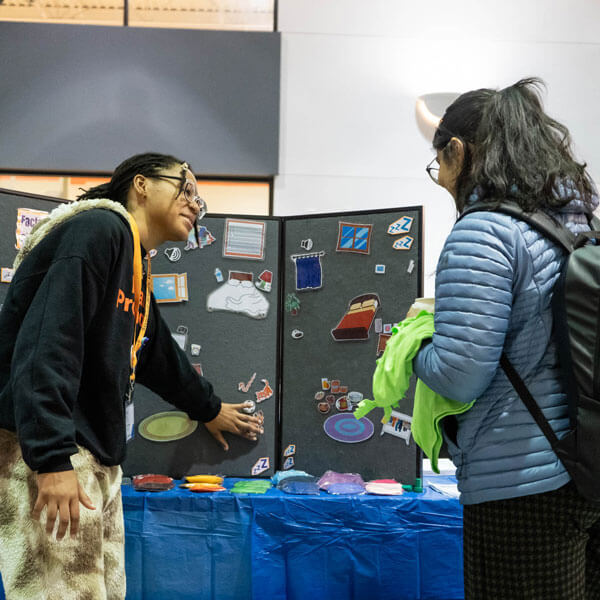 Two R I T students standing in front of a three panel board that has imagery of items related to sleep.