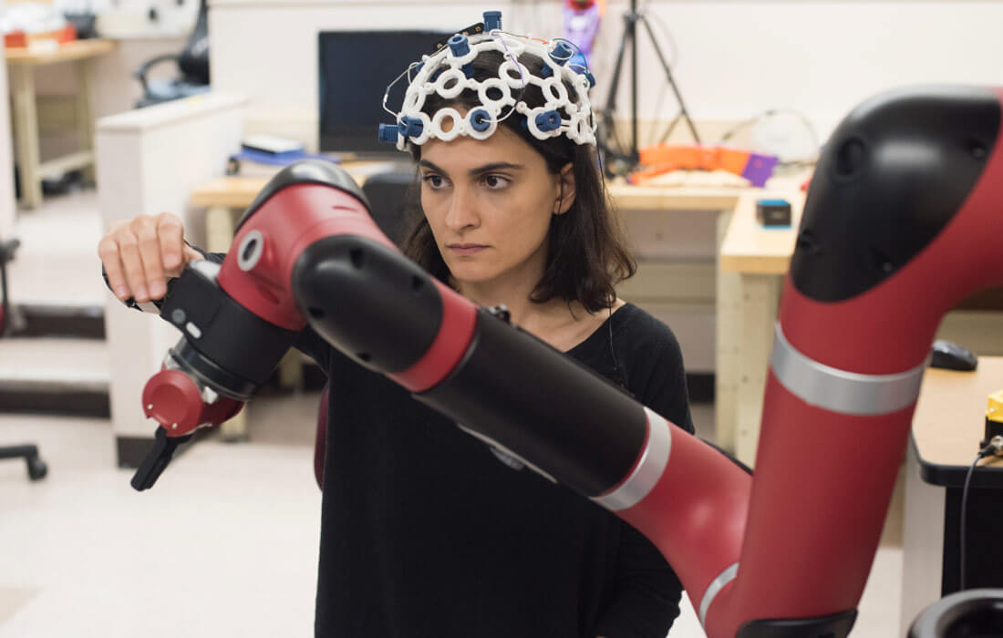 A female researcher wearing a helmet sensor rig, interacting with a robotic arm.
