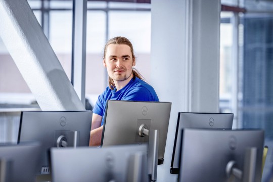 Man in blue shirt sitting among computer monitors, looking at the camera.