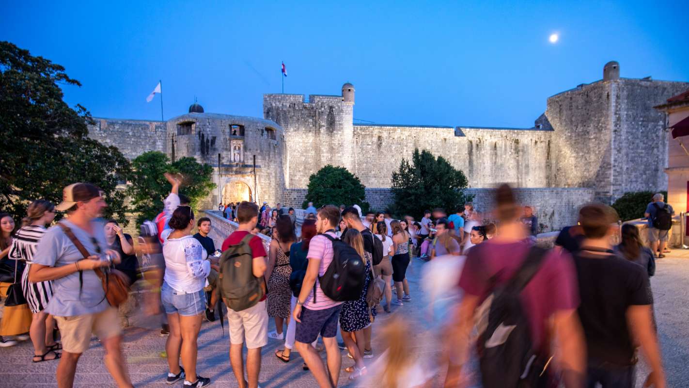 A crowd of people gathered outside Dubrovnik's historic city walls in the evening, illuminated by soft twilight and a glowing moon.