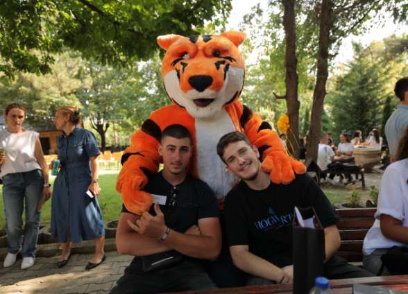 Two students pose with a person dressed as an orange tiger mascot, surrounded by trees and outdoor seating.